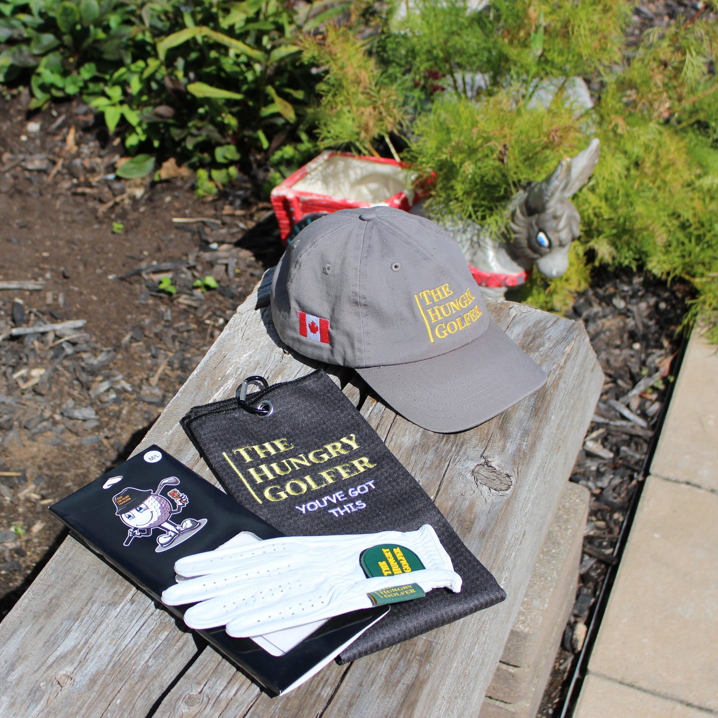 Golf hat, leather glove, and golf towel, all with the Hungry Golfer logo, sitting on a wooden block in a garden.