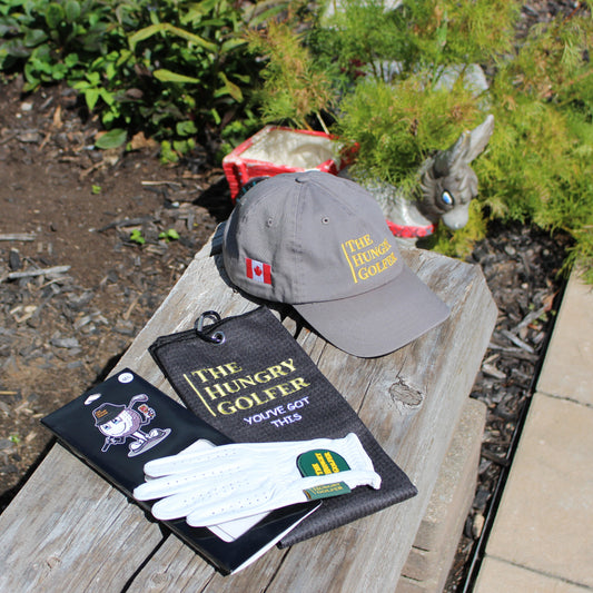 A golf cap, a leather golf glove, and a golf towel, all wothhh the Hungry Golfer logo, sitting on a log bench in a garden.